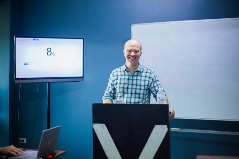 Smiling man giving a lecture at a podium with a whiteboard and monitor in a classroom.